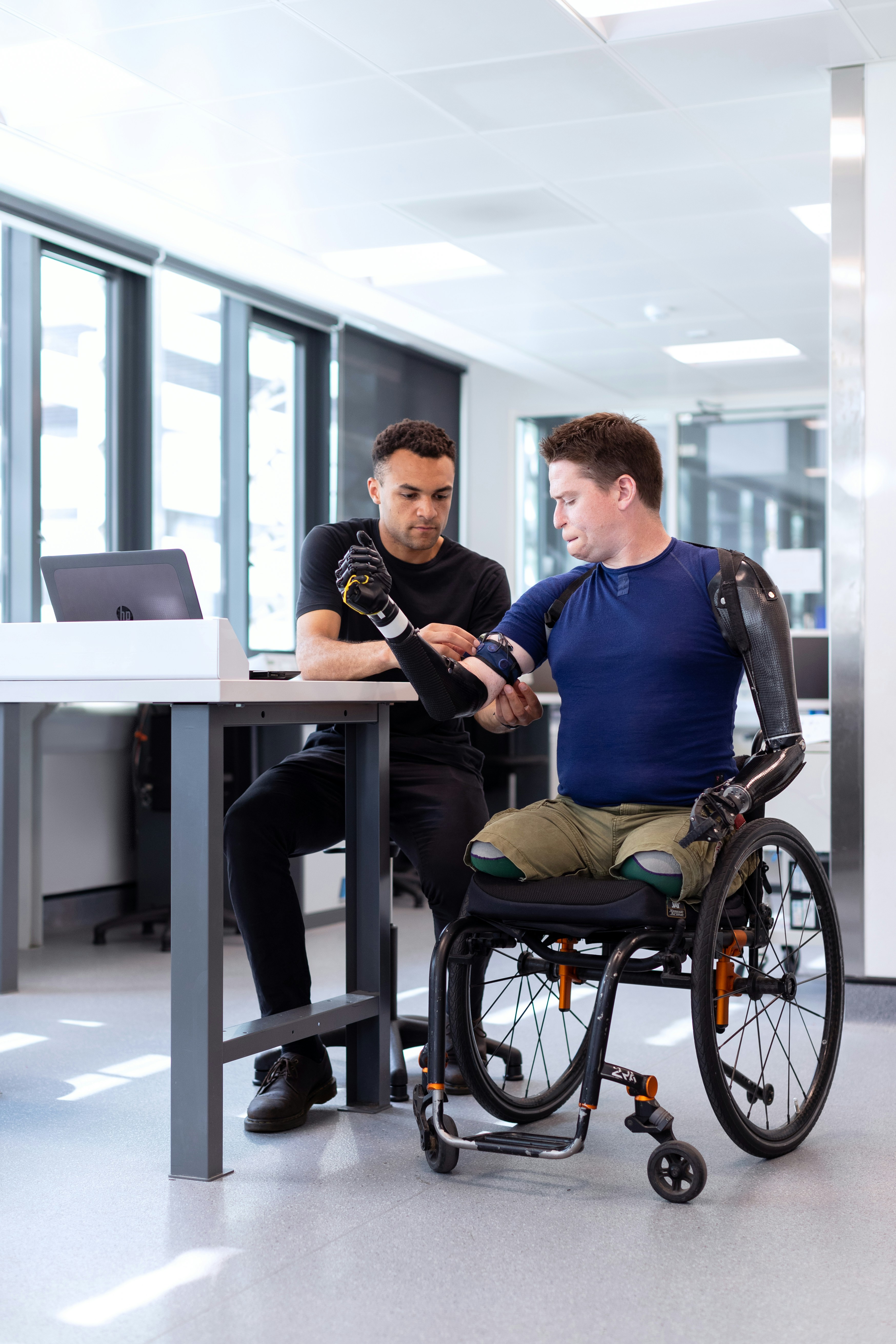 Man in wheelchair with prosthetic arms, assisted by another man at a computer